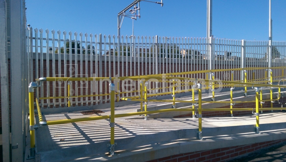 Close-up of Interclamp Key Clamp DDA Assist fittings and safety yellow handrail tube forming a secure and visible handrail on a disabled access ramp at a train station.